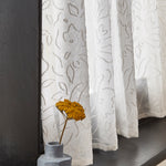 A window sill with a vase and wildflower against a sheer curtain in a hand drawn floral pattern in ivory.