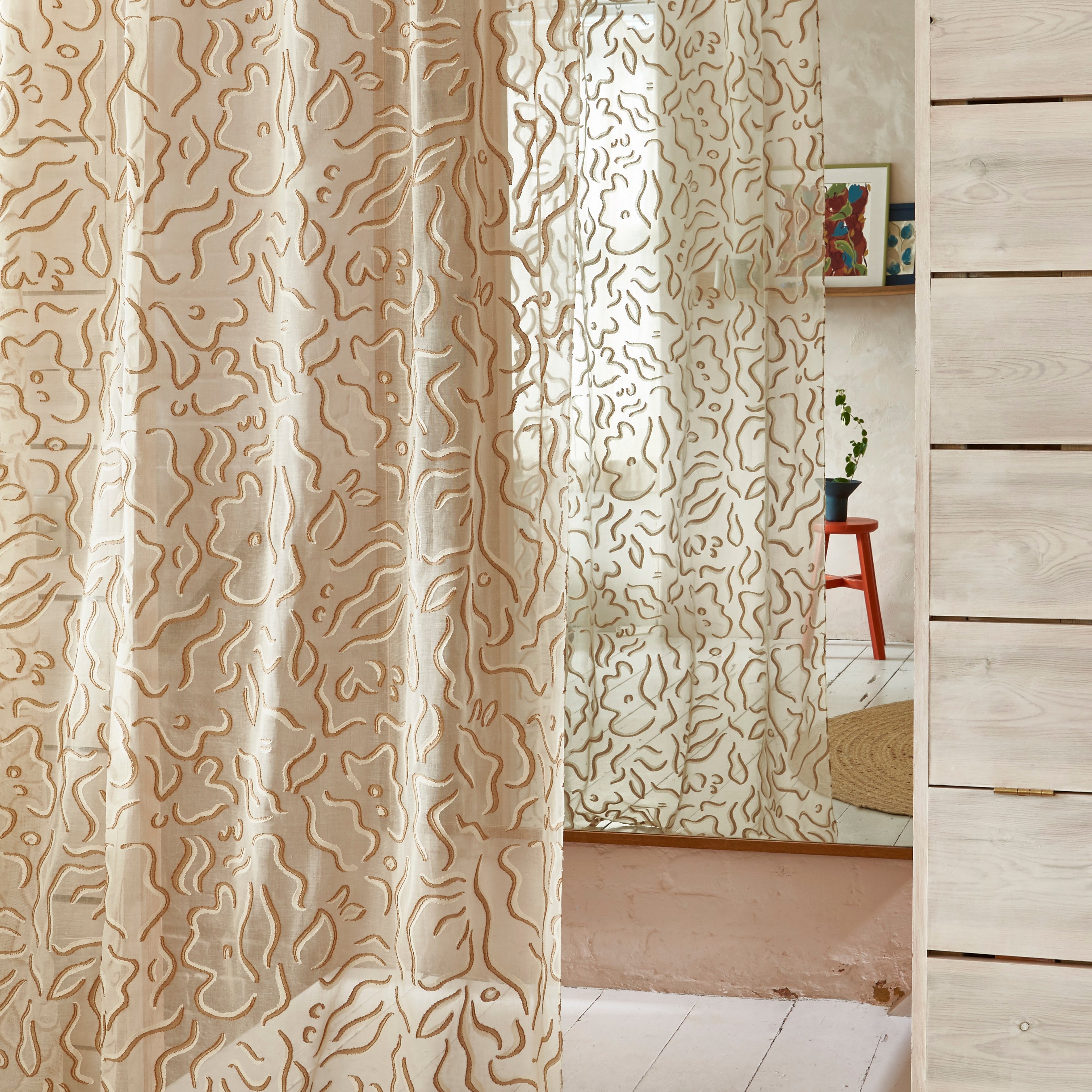Draped sheer floral patterned fabric against a mirror reflecting a rustic cabin interior with white washed wood, a red stool and a picture rail with framed paintings. 