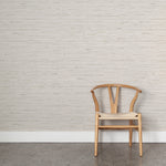 Wooden chair with woven seat on concrete floor against wallpaper in a textural striped pattern in shades of beige and grey.
