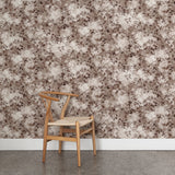 Angled wooden chair with woven seat on concrete floor against wallpaper in a painterly leaf print in shades of warm wine purple with a mottled white overlay.