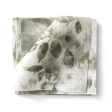 Stacked swatches of fabric in a painterly leaf print in shades of muted grey-green on a white background.