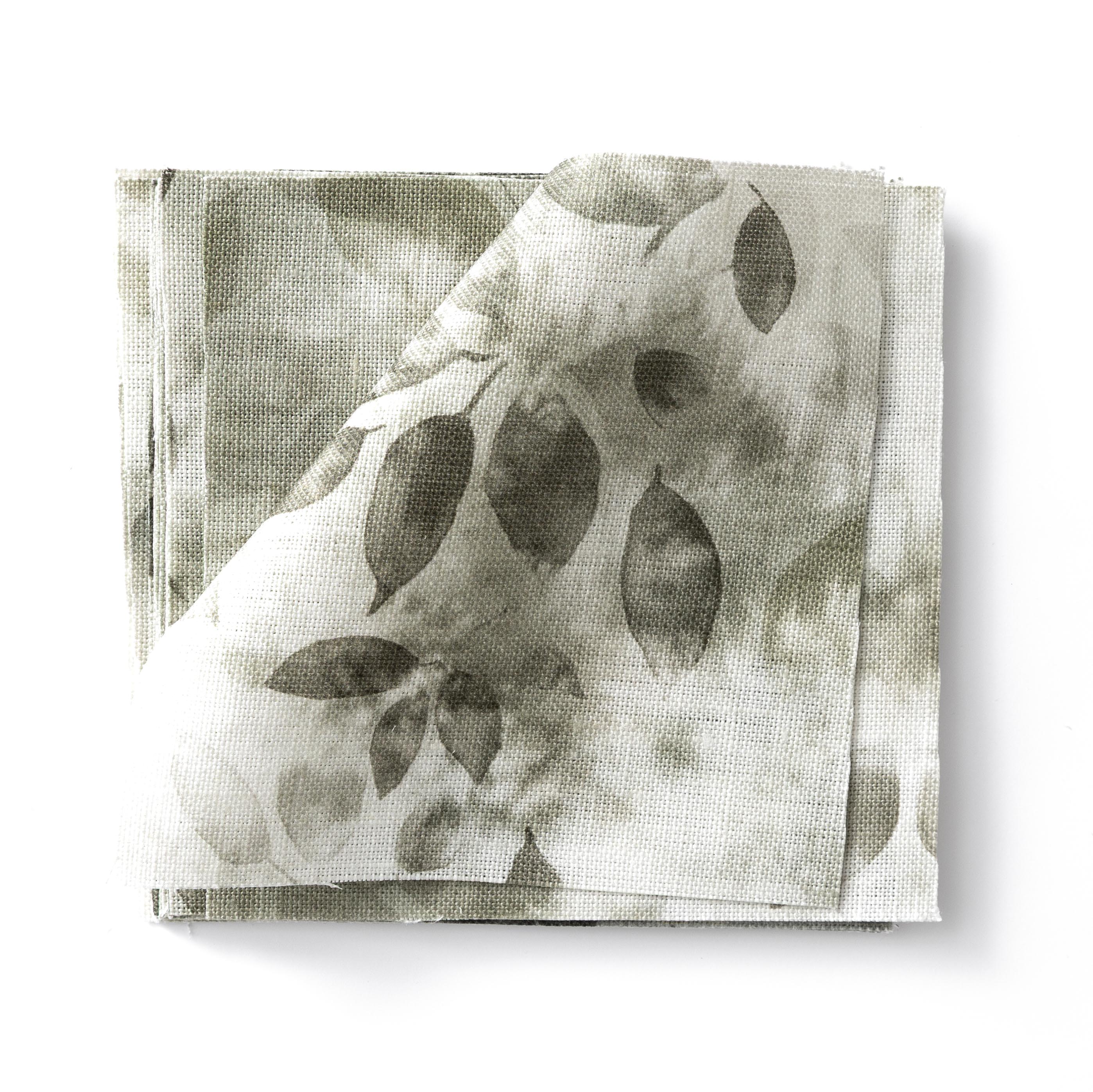 Stacked swatches of fabric in a painterly leaf print in shades of muted grey-green on a white background.