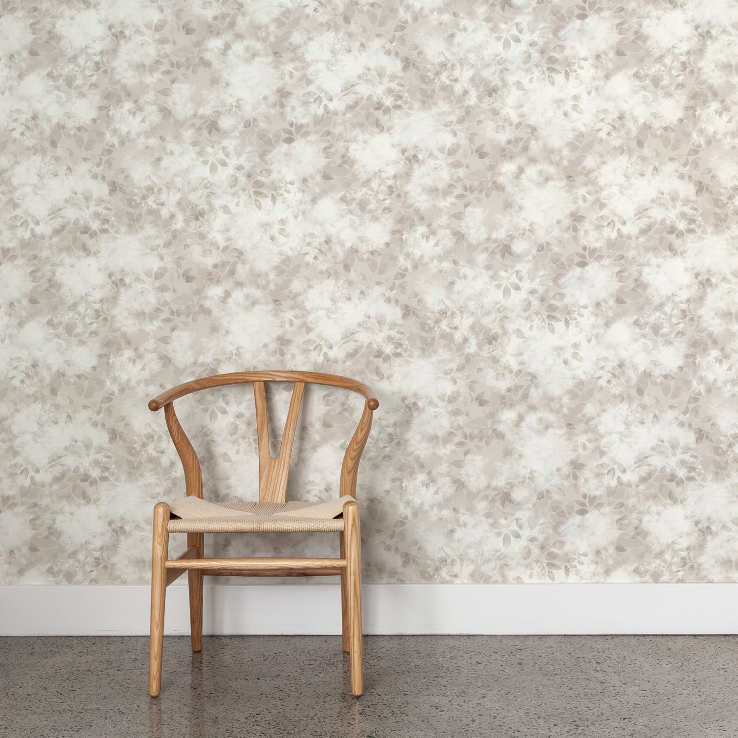 Wooden chair with woven seat on concrete floor against wallpaper in a painterly leaf print in shades of pinky grey with a mottled white overlay.