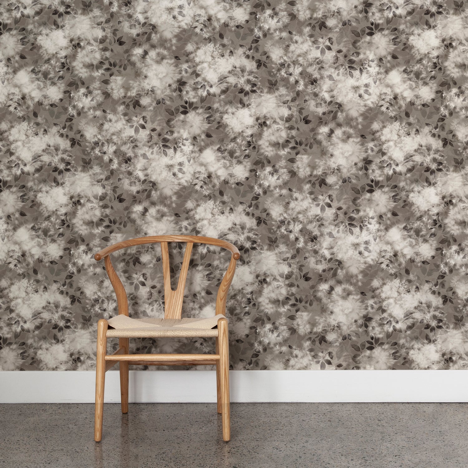 Angled wooden chair with woven seat on concrete floor against wallpaper in a painterly leaf print in shades of warm charcoal grey with a mottled white overlay.