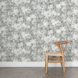Angled wooden chair with woven seat on concrete floor against wallpaper in a painterly leaf print in shades of cool steel grey with a mottled white overlay.