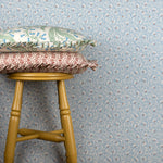 Wooden stool with decorative pillows against wallpaper in a lyrical wildflower design in white with red accents on a blue field. 