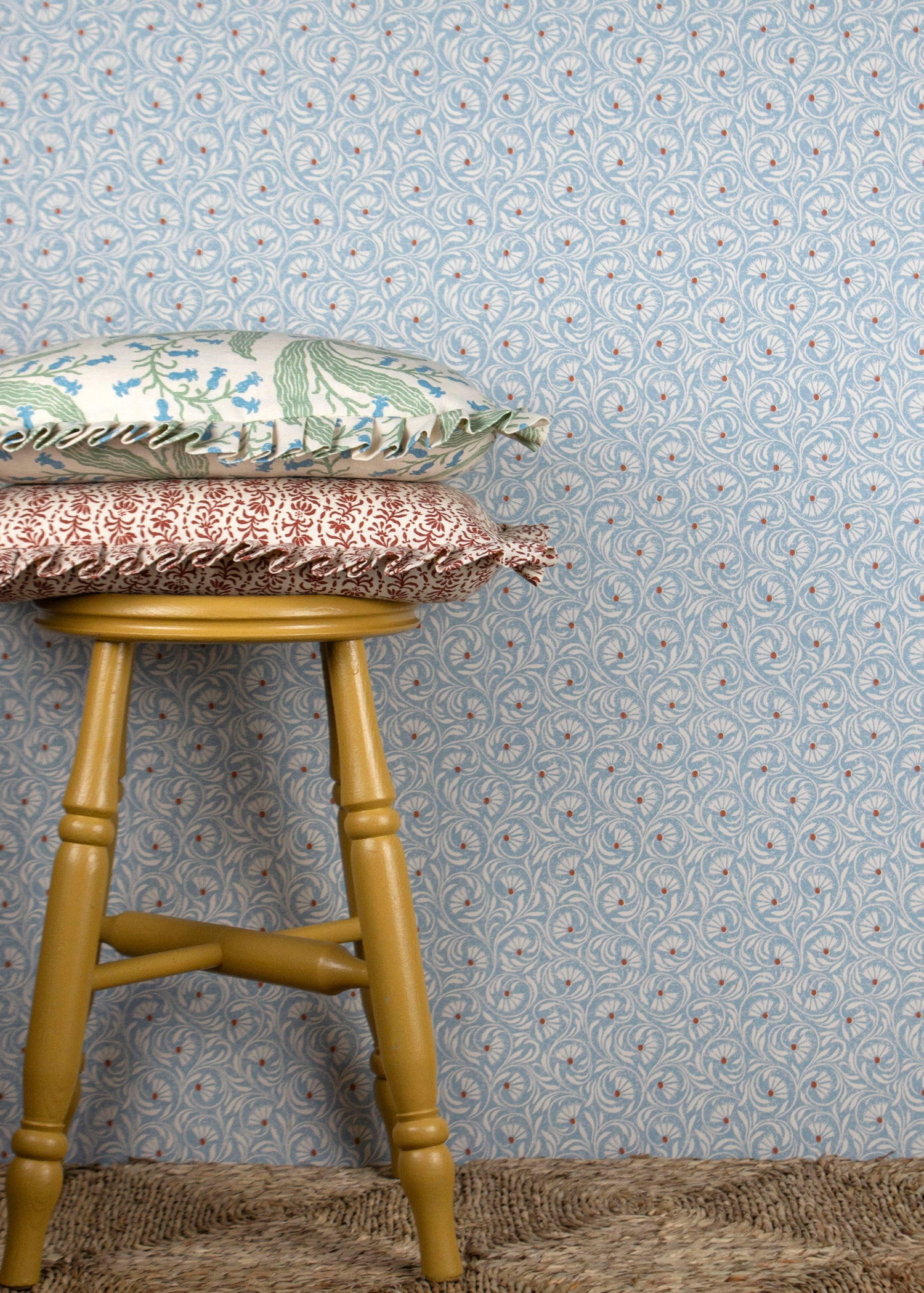 Wooden stool with decorative pillows against wallpaper in a lyrical wildflower design in white with red accents on a blue field. 