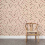 A wooden chair stands in front of a wall papered in a minimal repeating leaf print in pink on a tan field.