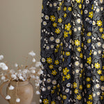 A vase of flowers stands in front of a draped curtain in a small-scale floral print in white, gray and yellow.