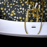 Detail shot of gilded bathtub faucets in front of a wall papered in a small-scale floral print in white, black and yellow.