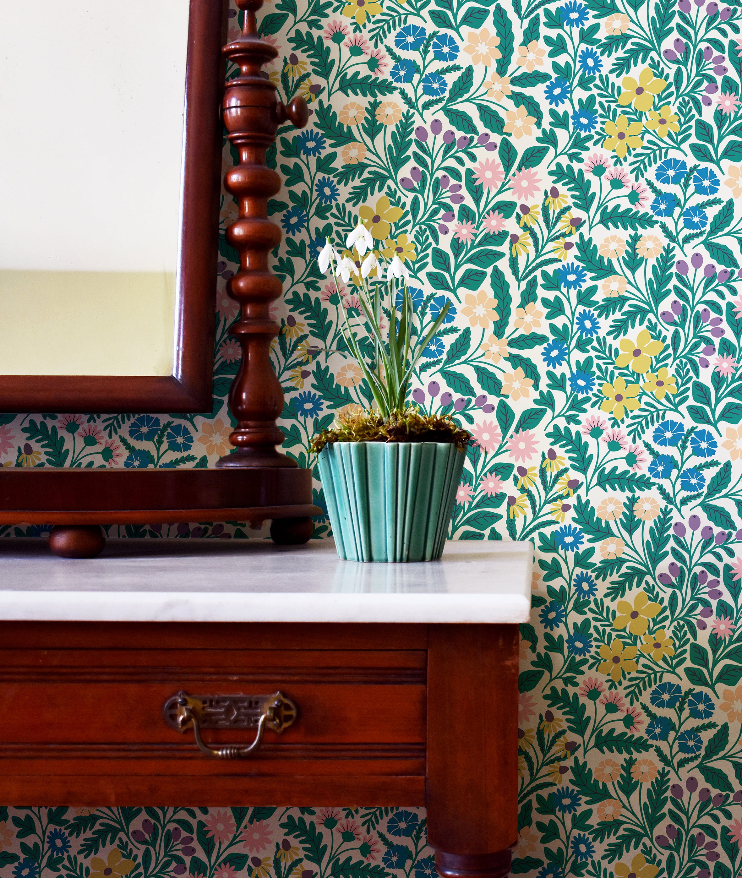 An end table with a potted plant stands in front of a wall papered in a multicolor small-scale floral print.