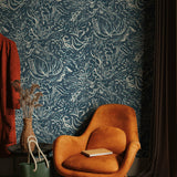 A burnt orange lounge chair in front of a wall papered in a painterly botanical print in white and blue on a navy field.