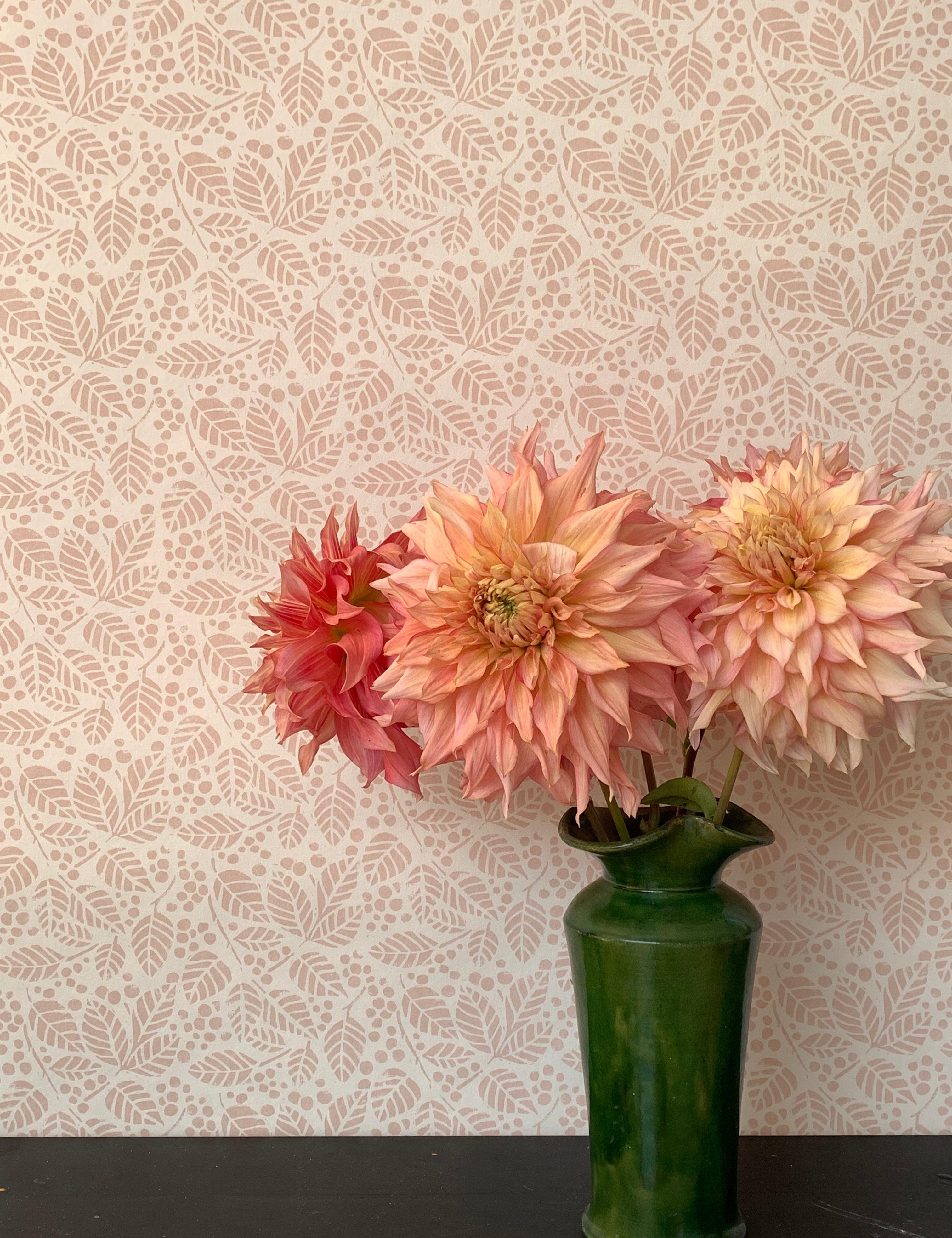 A green vase with pink peonies against a botanical leaf patterned wallpaper in soft pink.