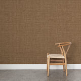 Angled wooden chair with woven seat on concrete floor against wallpaper in a textured tweed pattern in shades of warm caramel brown. 