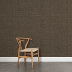 Angled wooden chair with woven seat on concrete floor against wallpaper in a textured tweed pattern in shades of warm charcoal grey.