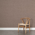 Angled wooden chair with woven seat on concrete floor against wallpaper in a textured tweed pattern in shades of muted mauve purple. 