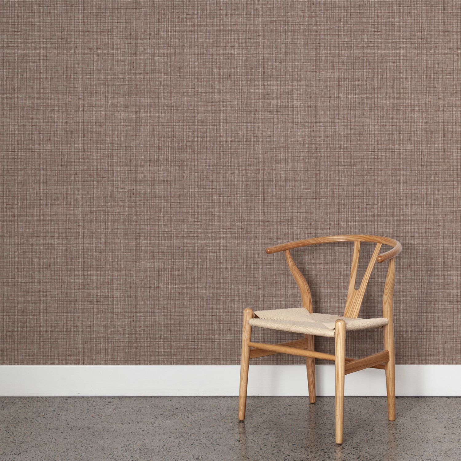 Angled wooden chair with woven seat on concrete floor against wallpaper in a textured tweed pattern in shades of muted mauve purple. 