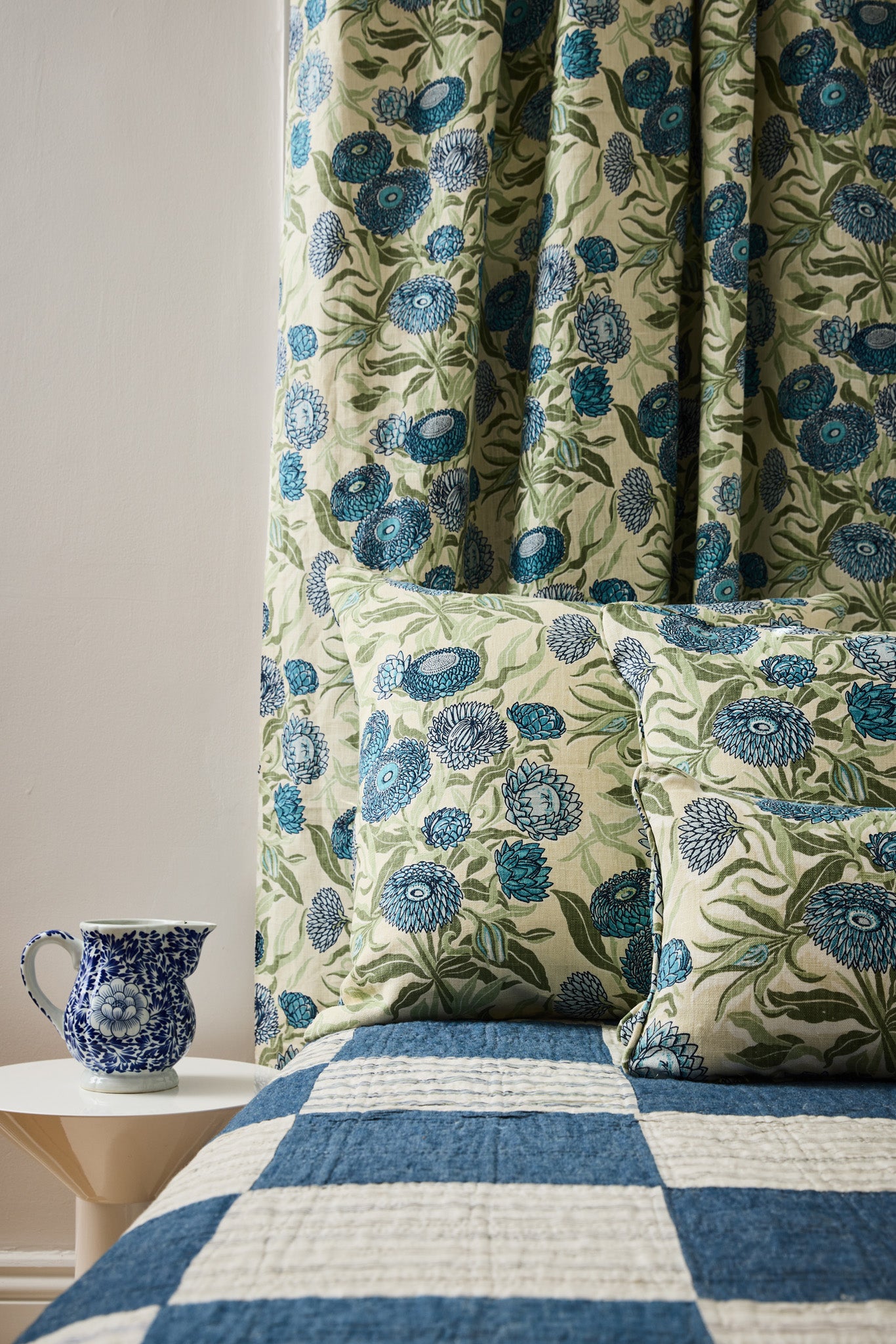 Bedrooms scene with checkerboard quility and pillows in a floral orinted linen in a daisy design in shades of blue with green leaves on a pale cream field.