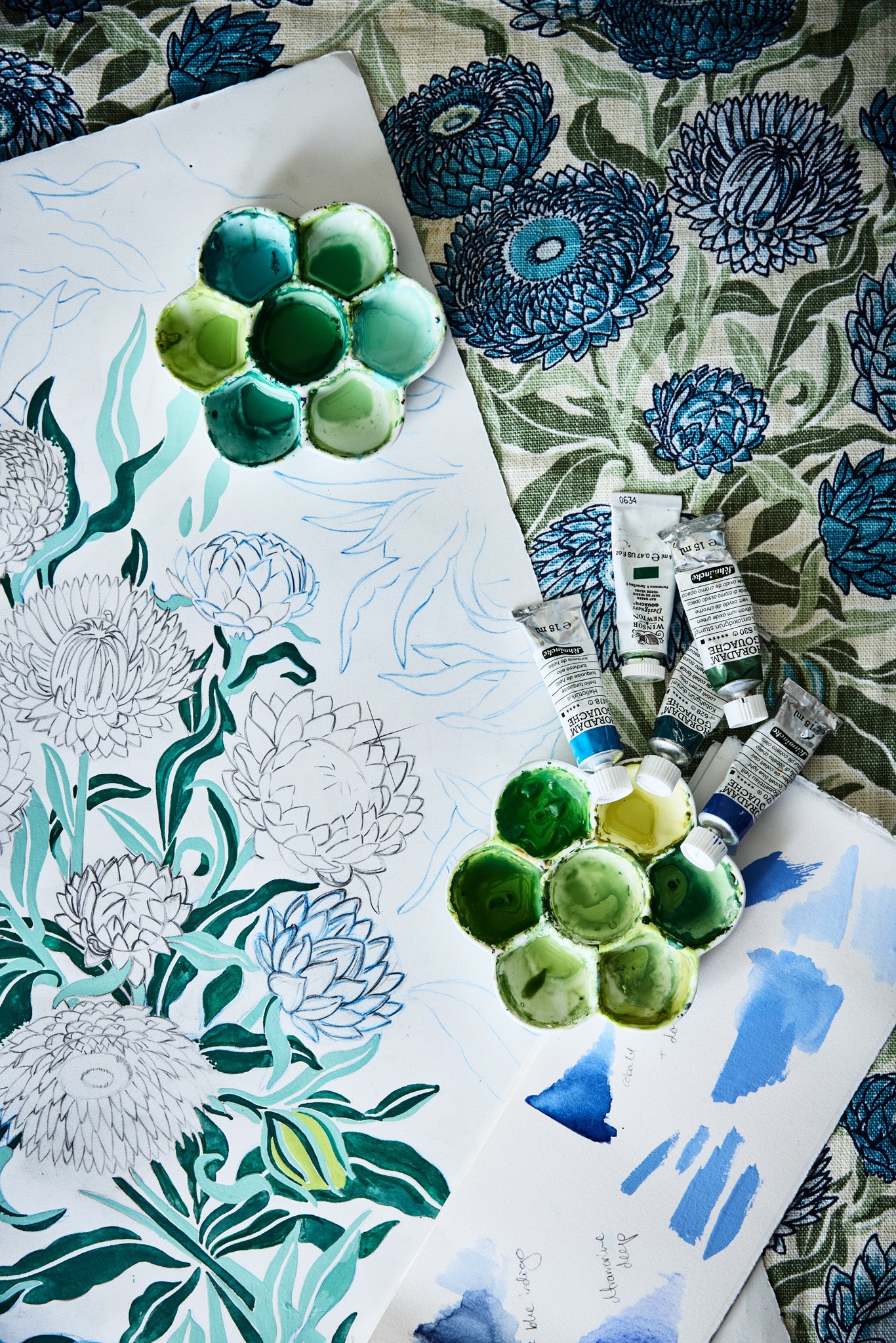 Overhead image of an artists process working on a botanical floral design n shades of blue and green with paint tubes, palettes and fabric background.