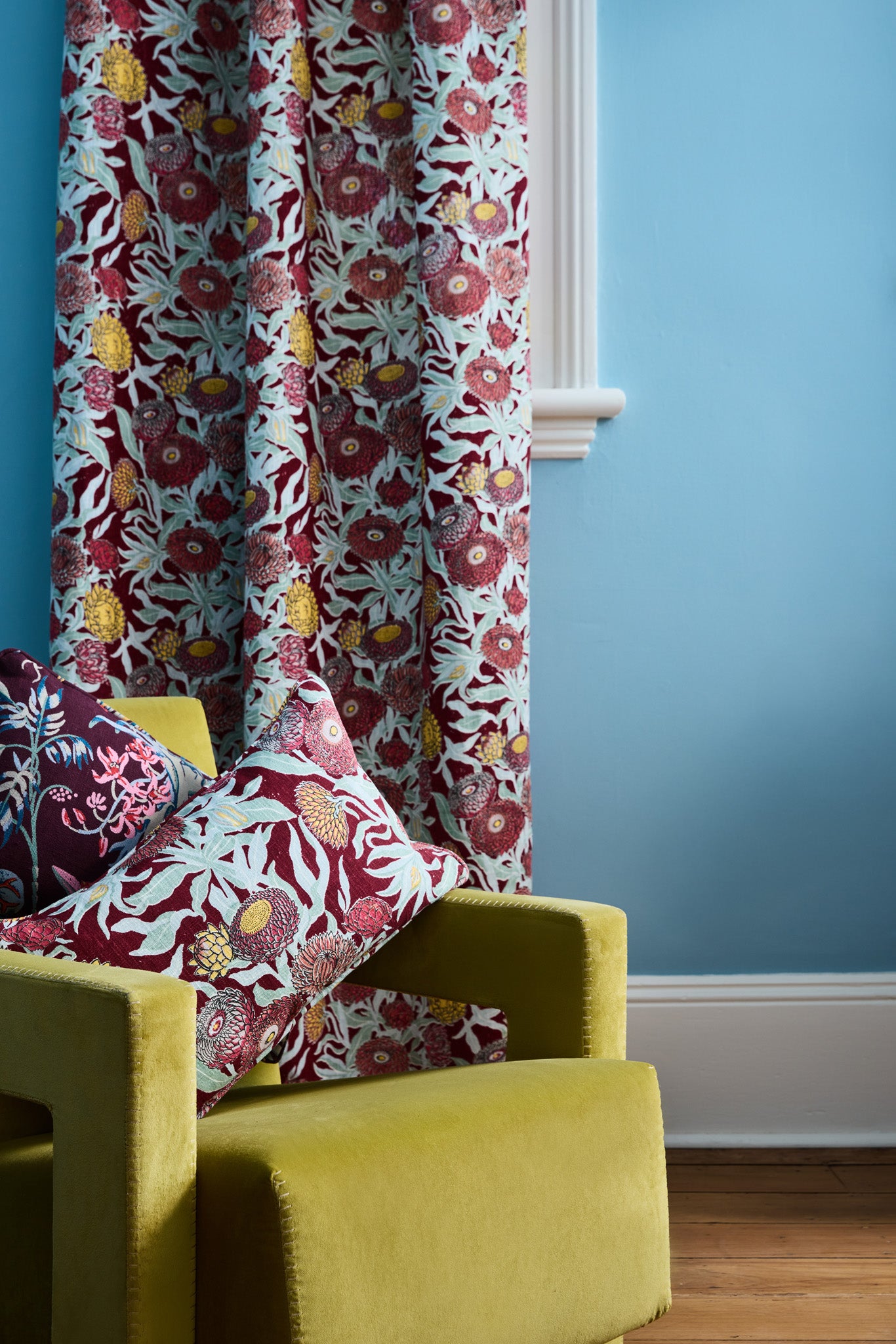 Lime green velvet chair with read and turquoise floral patterns against a matching curtain on a bright blue wall. 