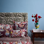Bedroom scene with a bedding in floral patterns in red, turquoise and green with a wooden side table with vase and flowers against a blue wall. 