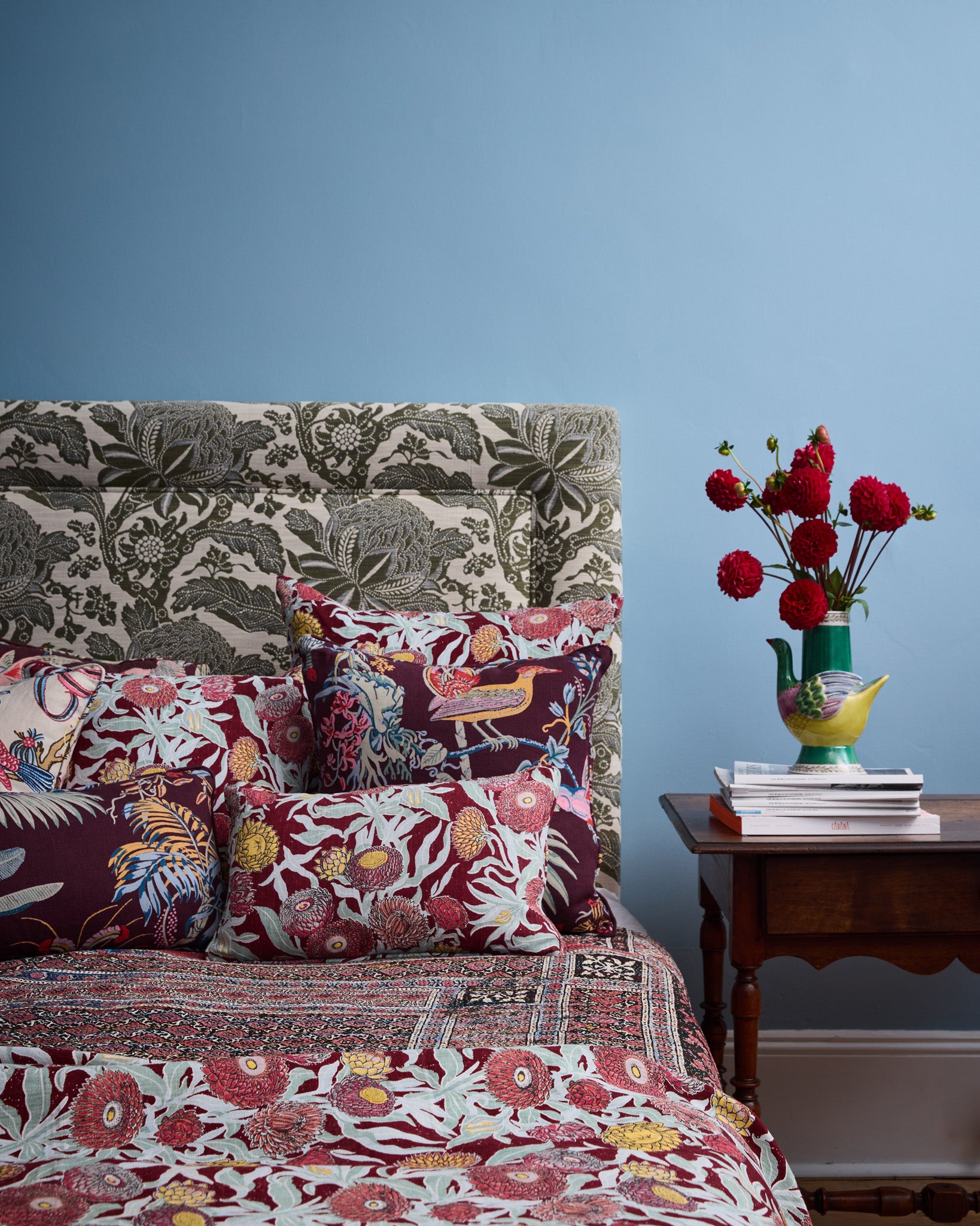 Bedroom scene with a bedding in floral patterns in red, turquoise and green with a wooden side table with vase and flowers against a blue wall. 
