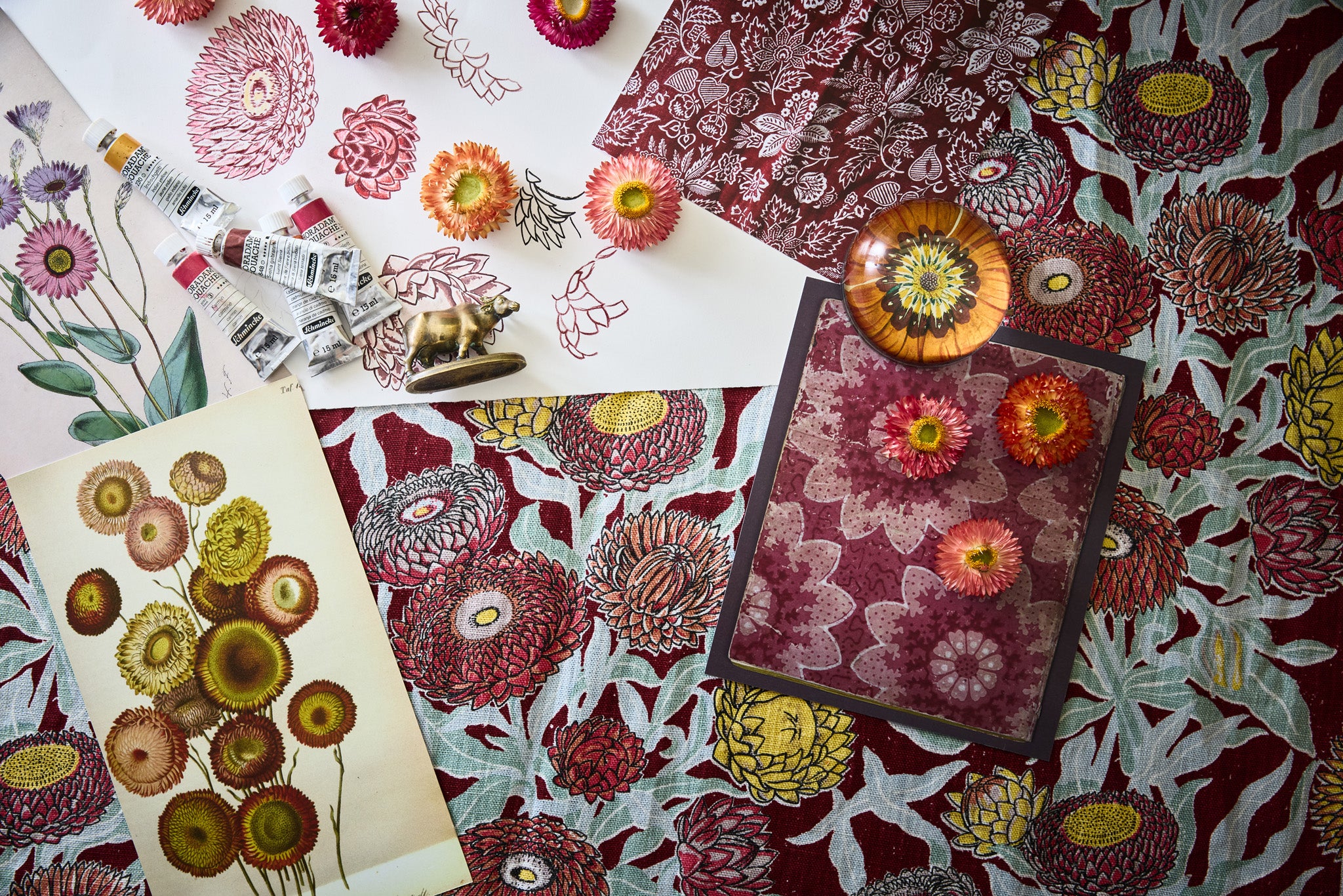 Overhead image of an artists process working on a botanical floral design n shades of red, yellow and aqua with paint tubes, fabric swatches and reference images.