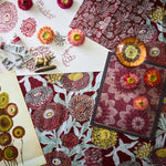 Overhead image of an artists process working on a botanical floral design n shades of red, yellow and aqua with paint tubes, fabric swatches and reference images.