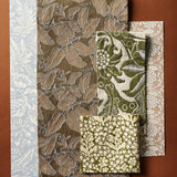 A flatlay with fabric and wallpaper swatches with botanical and floral patterns in muted tones of blue, brown, olive green, and beige, arranged on a solid brown background.