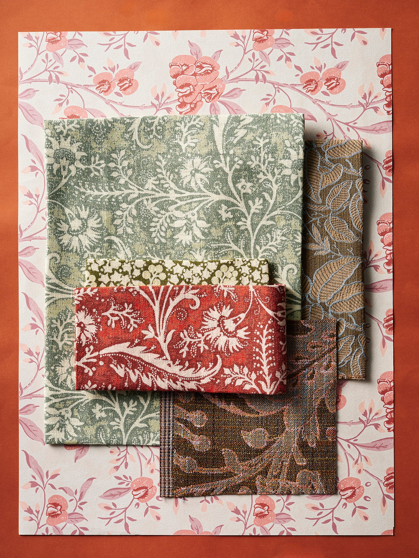 A mood board flat lay with patterned fabric in assorted botanical prints in shades of pink, brown, blush and red on a solid orange background.
