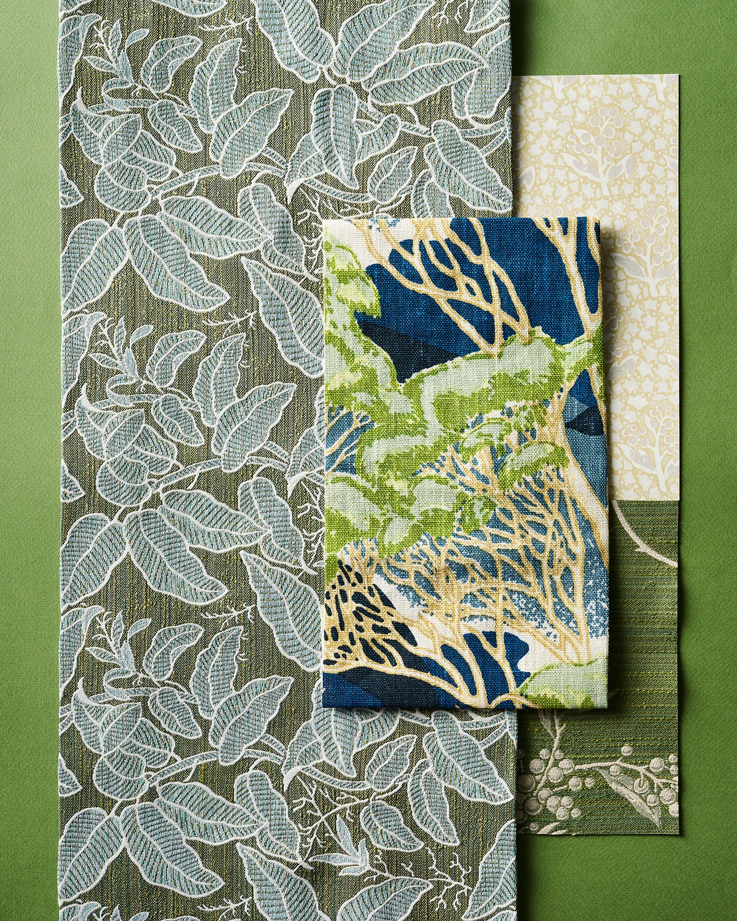 Flatlay with fabric swatches with botanical and leaf patterns in shades of blue, green, beige, and white, arranged on a solid green background.