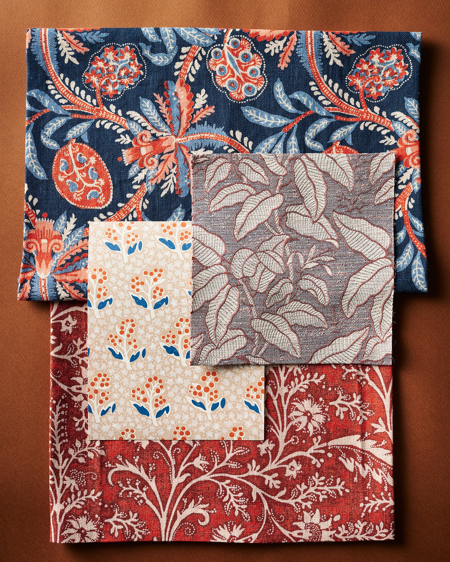 A flat lay with fabric and wallpaper swatches in botanical patterns in a color palette of blue, rust, red, and cream on a warm brown background. 