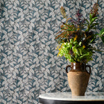 A white marble-topped table with a brown ceramic vase filled with a colorful assortment of foliage against a fabric backdrop with a botanical leaf pattern in grey and blue. 