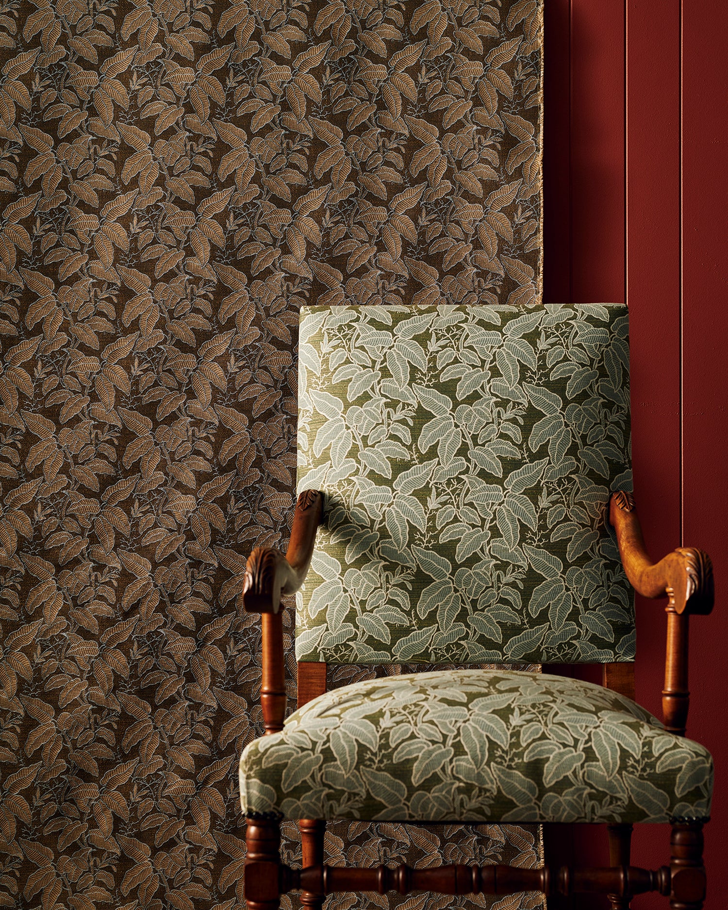 An upholstered wooden armchair with a leaf-patterned fabric in muted green and beige, set against a brown botanical fabric backdrop and a dark red wall panel.