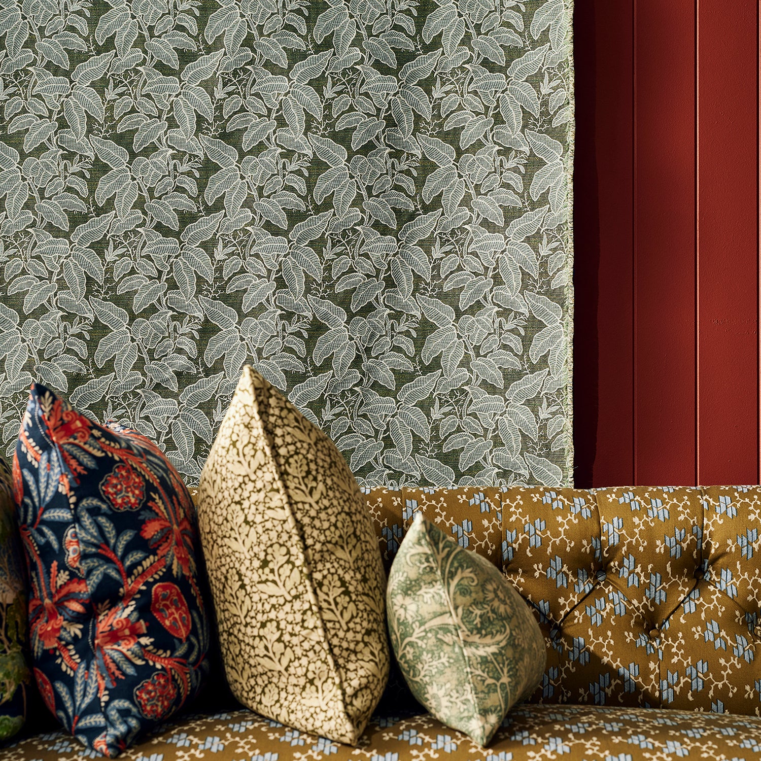Three decorative throw pillows in floral and botanical patterns arranged on a tufted mustard-yellow sofa against a green leaf patterned fabric background.