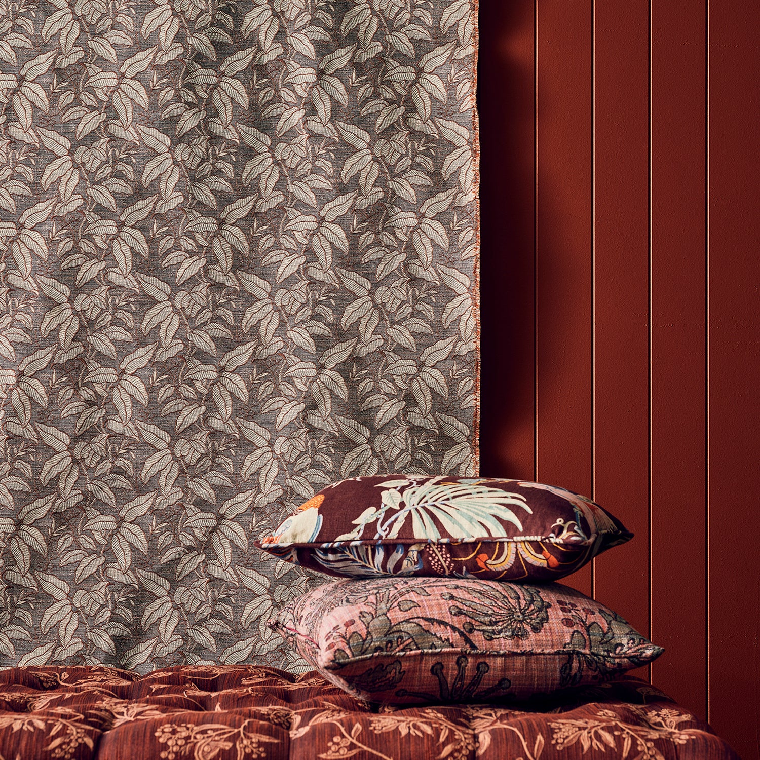 A styled interior scene showing patterned home textiles: a leafy curtain in taupe and brown, two decorative pillows with floral and ornamental prints, and a tufted upholstered bench in deep reddish-brown against a dark red paneled wall.