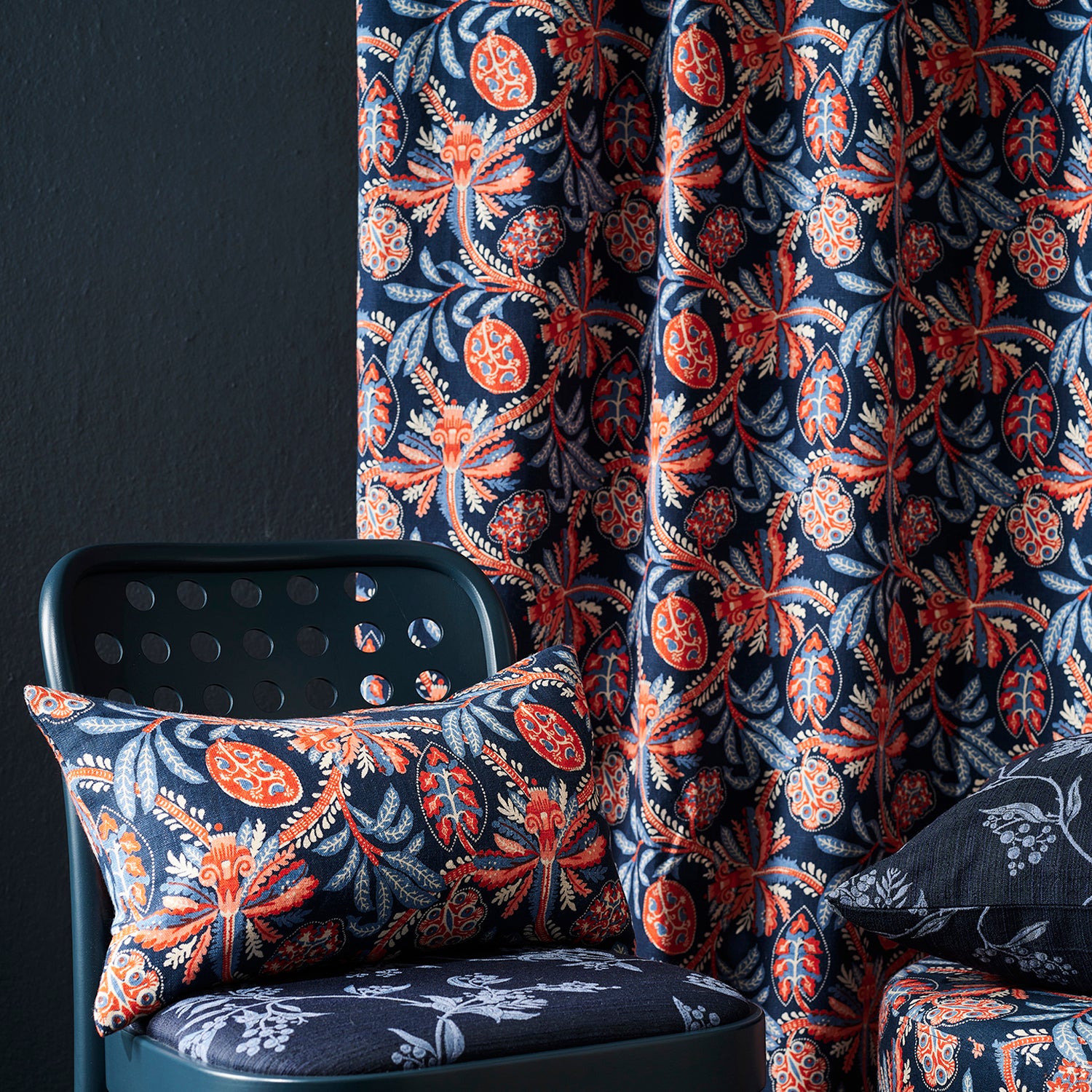 A close-up of botanical patterned fabric in a room setting, showing a curtain and matching cushion cover on a dark teal chair against a dark wall.