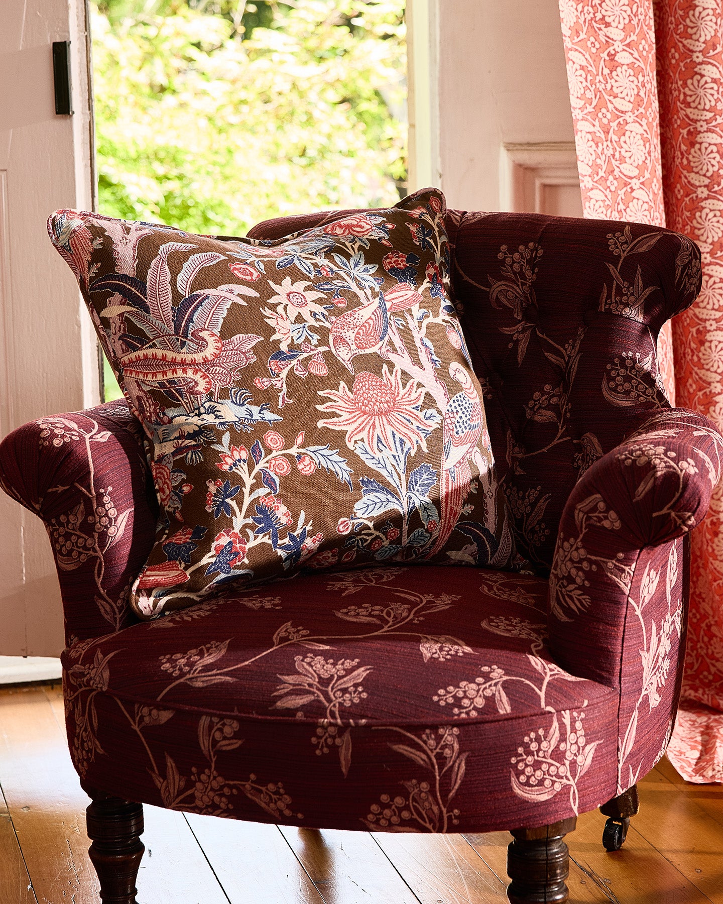 Patterned armchair with a floral pillow in a room with a window and curtains.