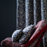 A close-up of botanical patterned curtains in navy blue and beige, with matching decorative pillows and a red-framed lounge chair in a dark room.