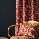 Red and beige scrolling vine patterned curtains and matching throw pillows styled in a room setting next to a wooden armchair.
