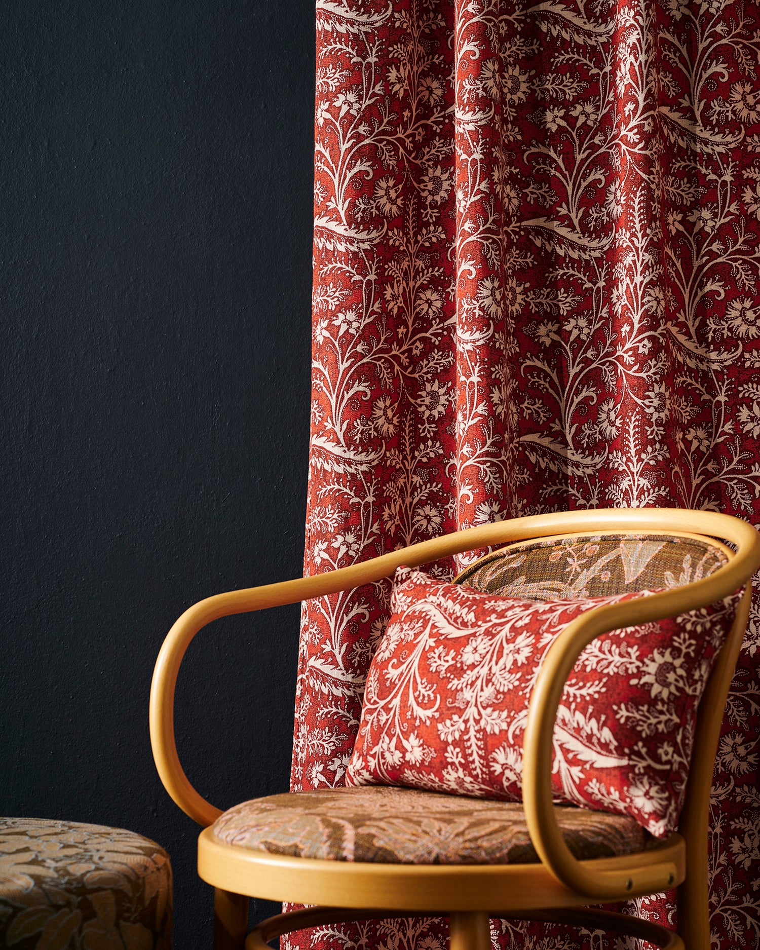 Red and beige scrolling vine patterned curtains and matching throw pillows styled in a room setting next to a wooden armchair.