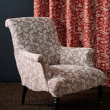 A classic wingback armchair upholstered in a warm grey and cream botanical leaf patterned fabric, set against a dark blue wall and a red floral patterned curtain.