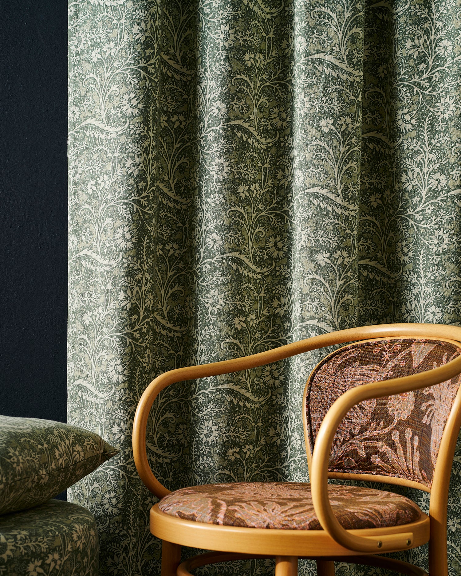 A close-up of a room featuring a green and beige botanical patterned curtain, a matching patterned cushion, and a wooden armchair with brown patterned upholstery.