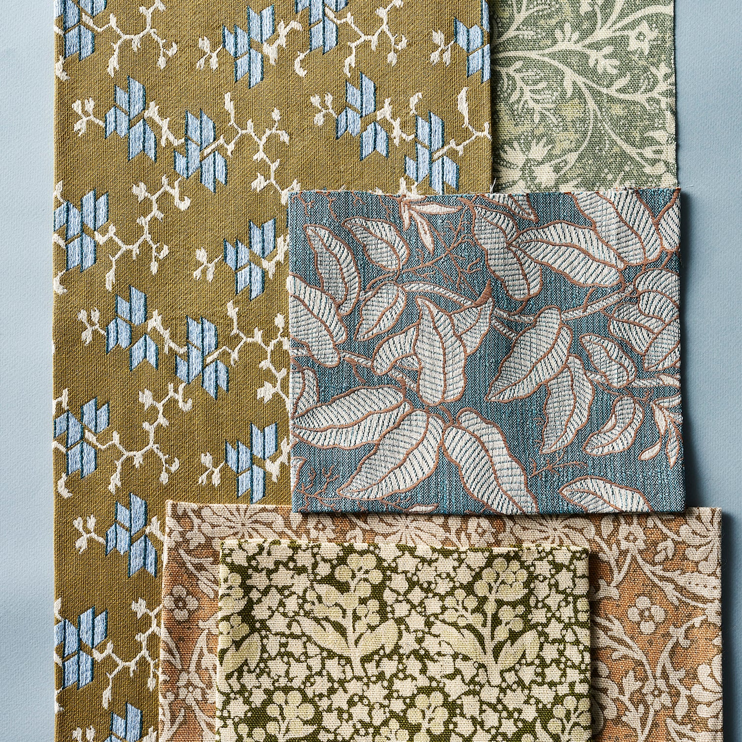 A set of woven fabric swatches with various botanical and geometric patterns, including olive green, dusty blue, and beige designs, arranged on a cool blue background.