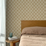 Bed with tan linens and wooden headboard, wooden side table  with white book and green glass, white floor length curtain against a watercolor checkerboard wallpaper in tonal warm tan. 