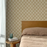 Bed with tan linens and wooden headboard, wooden side table  with white book and green glass, white floor length curtain against a watercolor checkerboard wallpaper in tonal warm tan. 