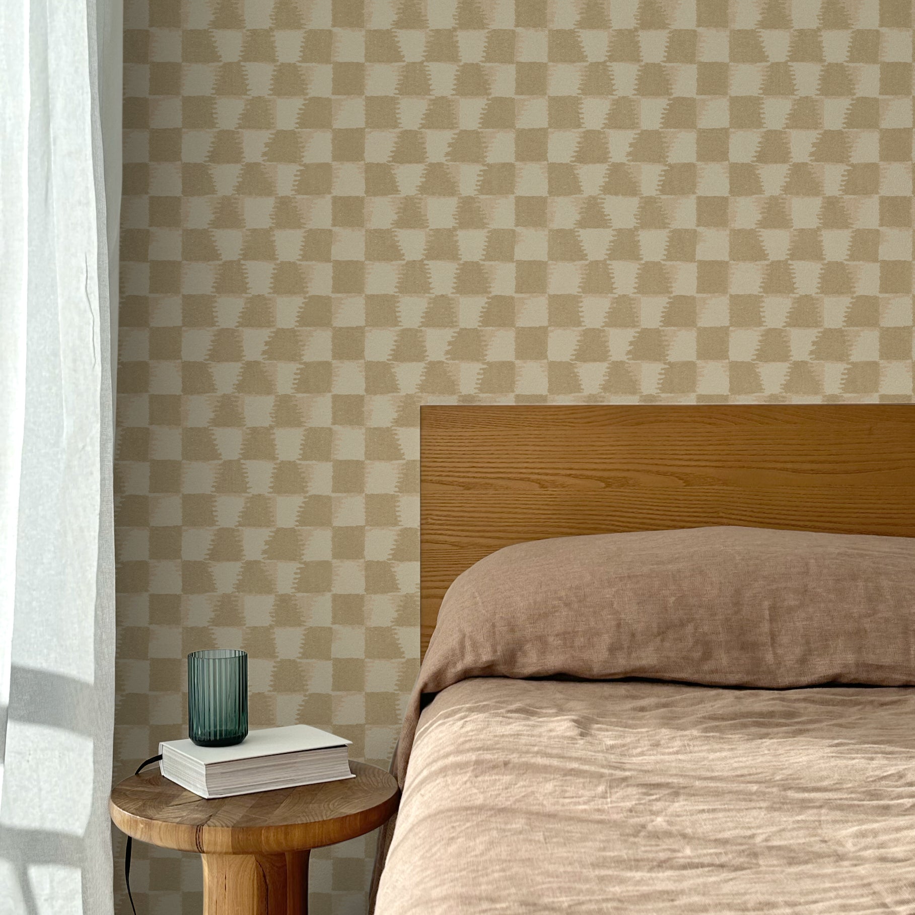 Bed with tan linens and wooden headboard, wooden side table  with white book and green glass, white floor length curtain against a watercolor checkerboard wallpaper in tonal warm tan. 