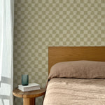 Bed with tan linens and wooden headboard, wooden side table  with white book and green glass, white floor length curtain against a watercolor checkerboard wallpaper in tonal sage green.