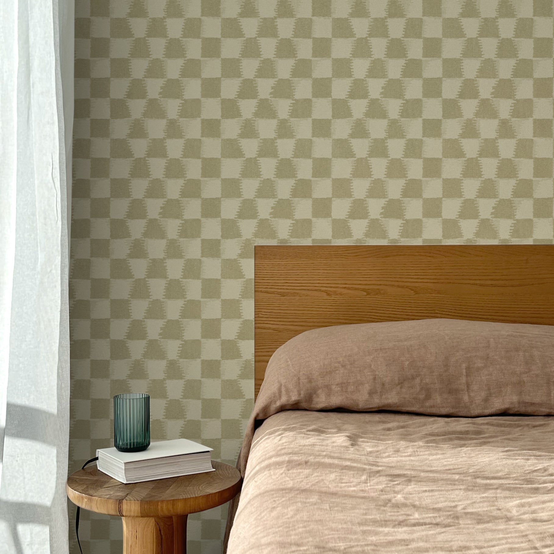 Bed with tan linens and wooden headboard, wooden side table  with white book and green glass, white floor length curtain against a watercolor checkerboard wallpaper in tonal sage green.