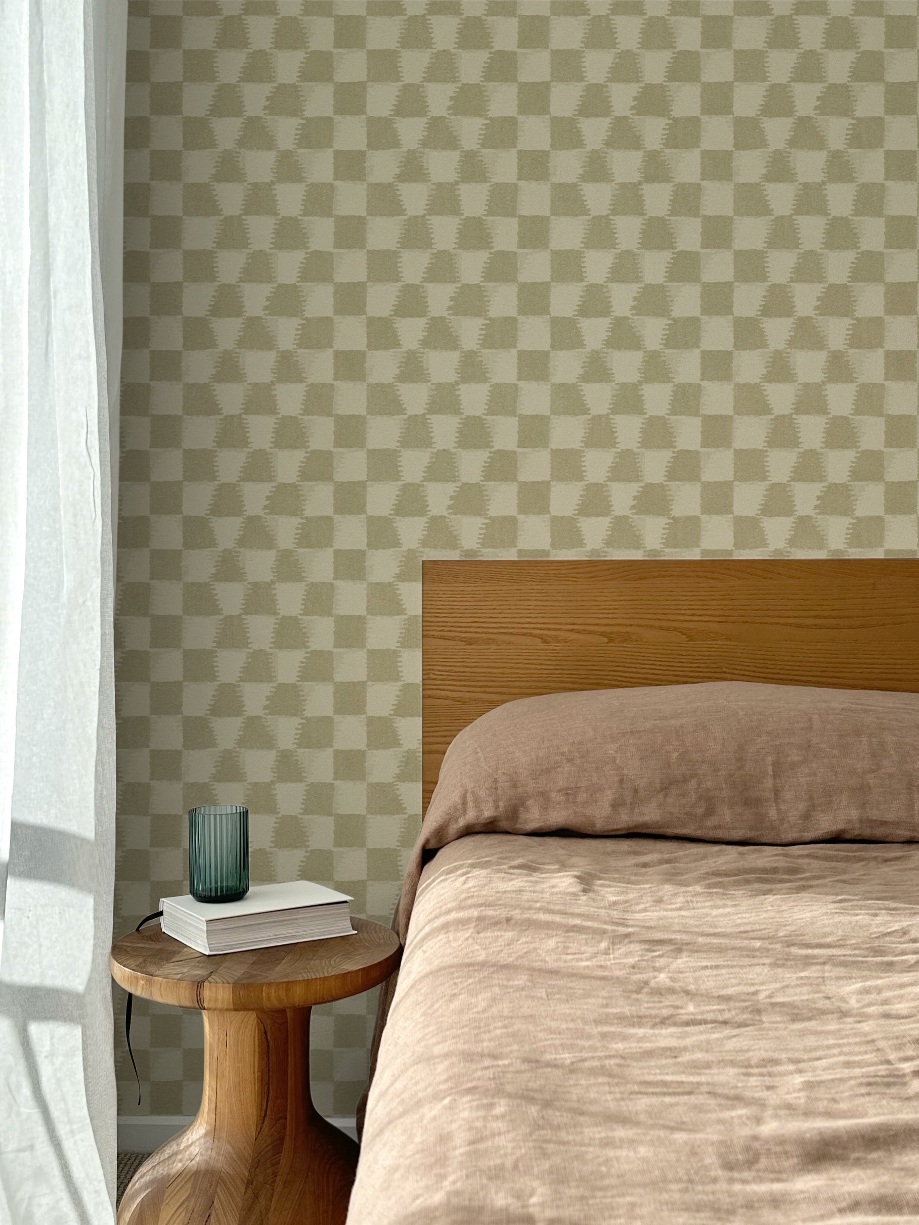 Bed with tan linens and wooden headboard, wooden side table  with white book and green glass, white floor length curtain against a watercolor checkerboard wallpaper in tonal sage green.